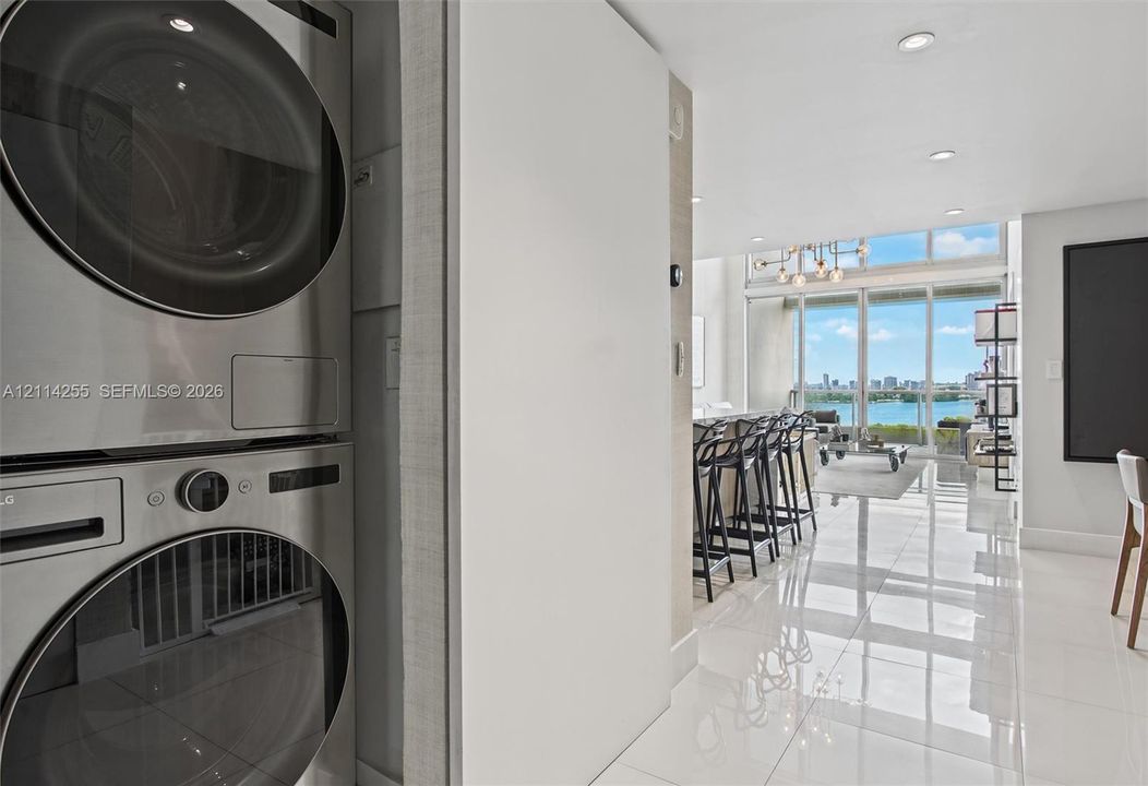 Stacked stainless steel laundry appliances, polished tile flooring, recessed ceiling lighting, and floor-to-ceiling glass paneling