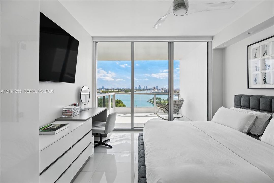 Floor-to-ceiling sliding glass doors, white tile flooring, recessed ceiling lighting, three-blade ceiling fan, and private balcony with water and city skyline backdrop