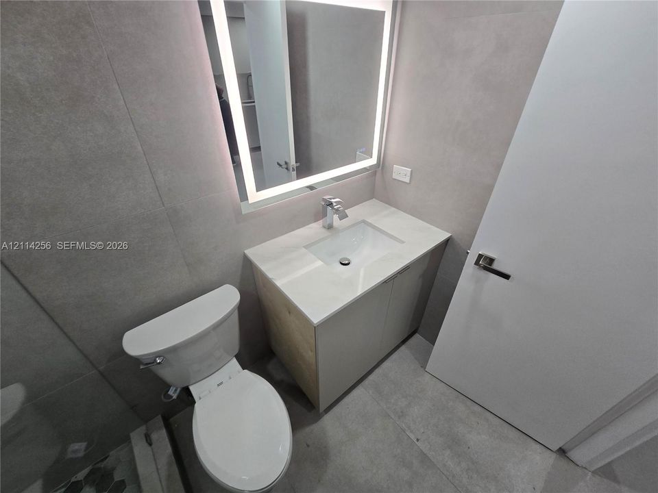 Modern vanity featuring a stone-composite countertop, integrated rectangular sink, and chrome faucet, paired with a LED-backlit mirror, large-format tile wall cladding, and a porcelain toilet