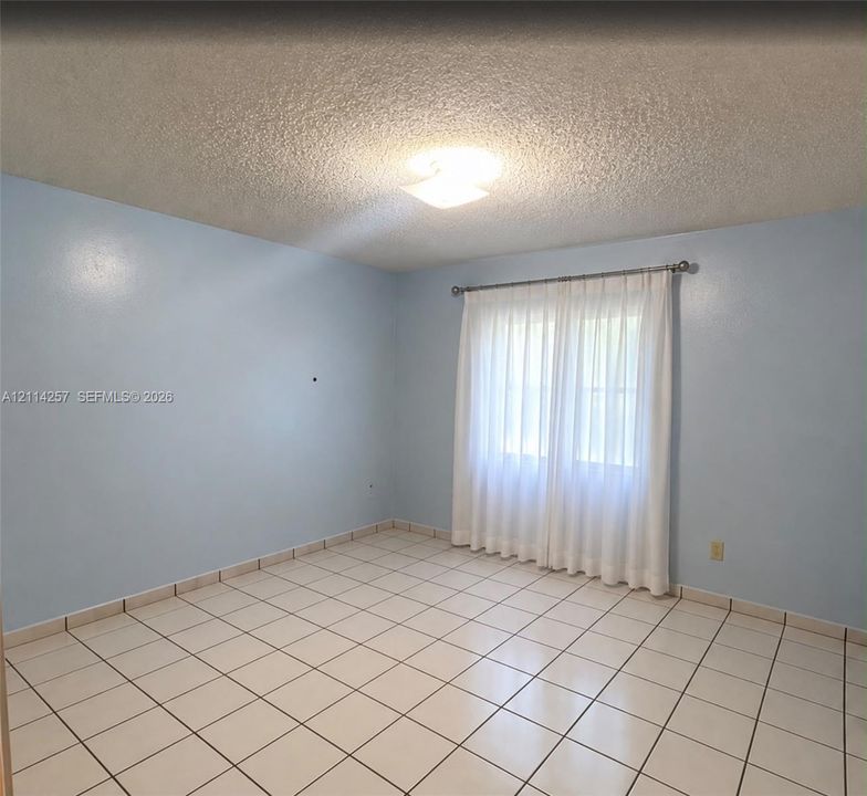 Room interior featuring light-toned ceramic tile flooring, solid walls, a centered overhead light fixture, and a window with a metal curtain rod