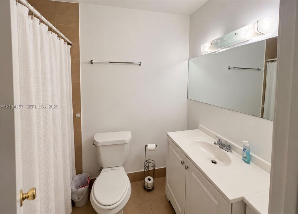 Bathroom featuring a white vanity cabinet with an integrated sink, full-width wall mirror, neutral tile flooring, and tub enclosure with tile surround