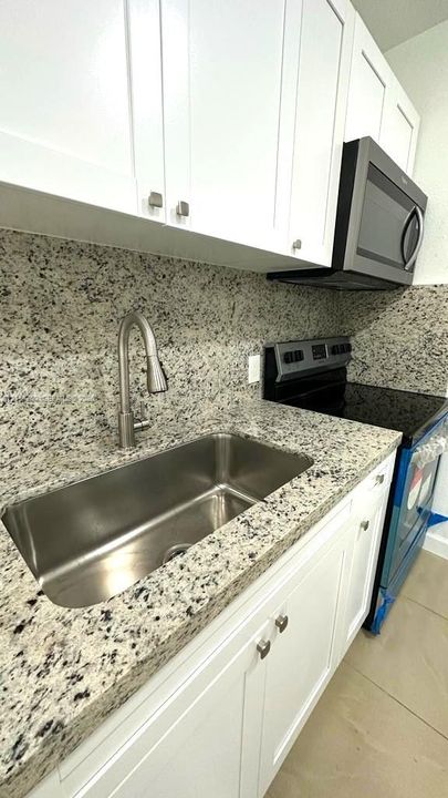 Kitchen featuring white shaker-style cabinetry, speckled stone countertops with matching backsplash, stainless steel under-mount sink with a high-arch pull-down faucet, and integrated stainless steel appliances