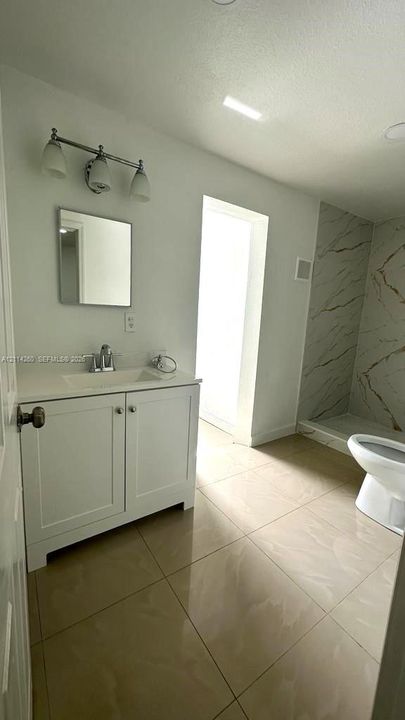 Bathroom featuring a white vanity cabinet with a stone-top integrated sink, polished chrome fixtures, a three-light vanity bar, and a walk-in shower with large-format stone-patterned wall tile and tile flooring