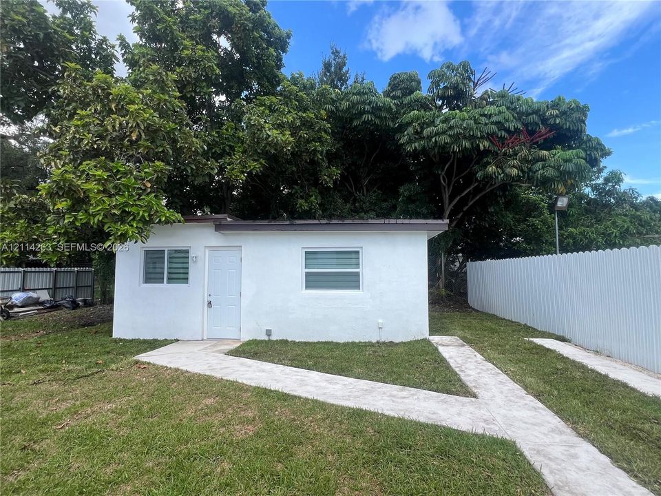 Detached structure featuring a white stucco exterior, metal roofing, dual horizontal sliding windows, and a paved walkway set within a fenced yard