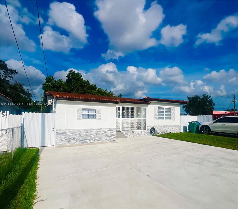 Single-story exterior featuring white stucco walls with stone-veneer wainscoting, a tiled roof, metal security bars on windows and entryway, and a concrete driveway