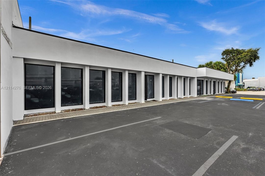 Modern exterior featuring a smooth stucco facade, expansive floor-to-ceiling windows with black framing, rhythmic architectural columns, and an asphalt-paved parking surface