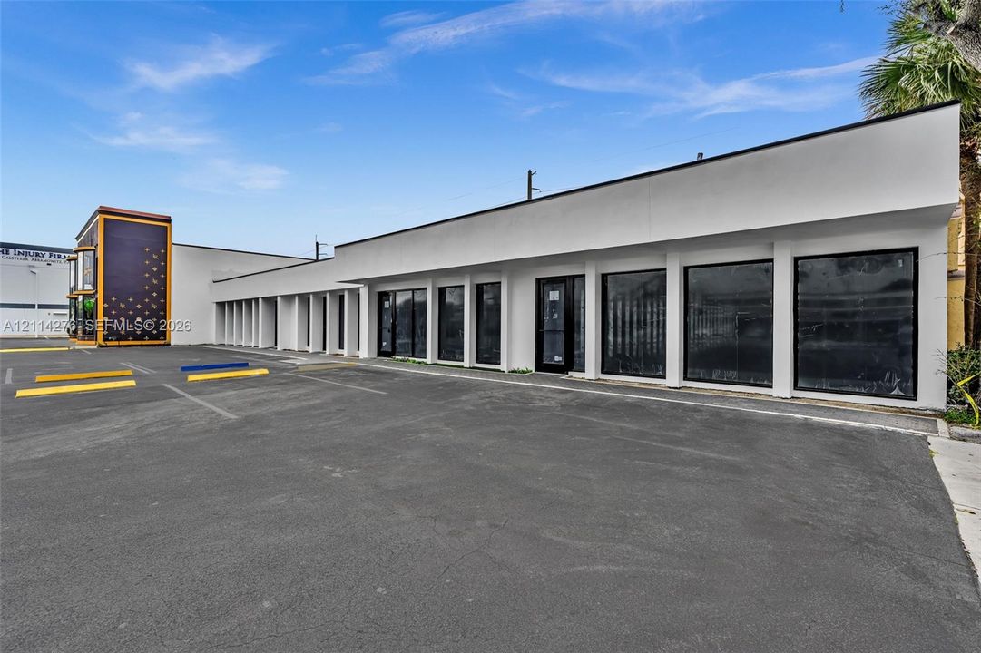 Commercial building exterior featuring a white smooth-finish facade, extensive floor-to-ceiling glass paneling, and an asphalt parking area with marked stalls