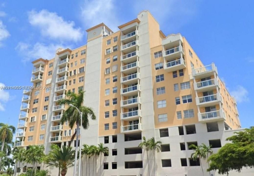 High-rise multi-story building featuring a stucco facade, stacked private balconies with glass railings, and ground-level covered parking