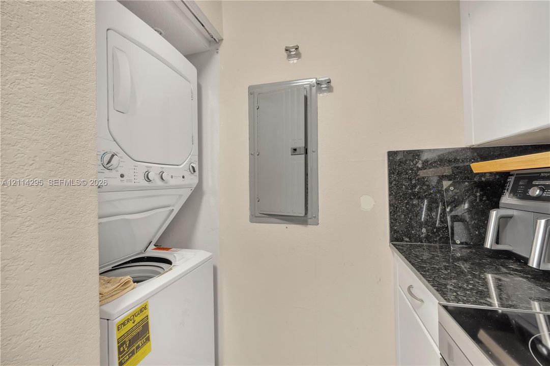 Stacked laundry unit, recessed electrical panel, white cabinetry, and stone-finish countertop