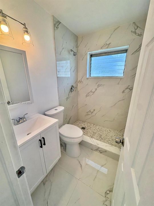 Bathroom featuring large-format veined tile flooring, white shaker-style vanity cabinet, walk-in shower with pebble-stone flooring, and dual-light vanity fixture