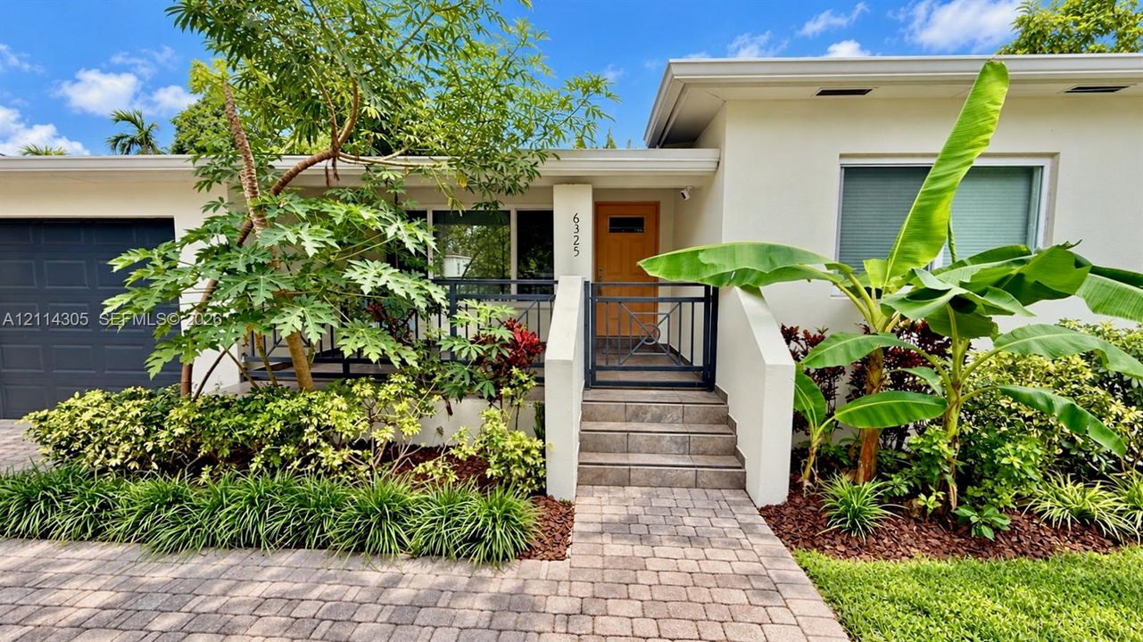 Stucco exterior featuring a paved stone walkway, covered front porch with metal railing, single-car garage, and mature landscaping