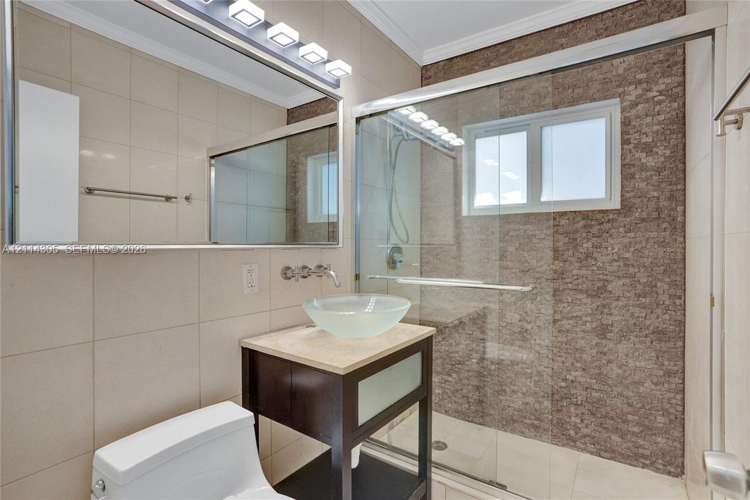Bathroom featuring a vessel sink, vanity with stone countertop, walk-in shower with a glass enclosure and stone-tile accent wall, and a large-format tiled wall