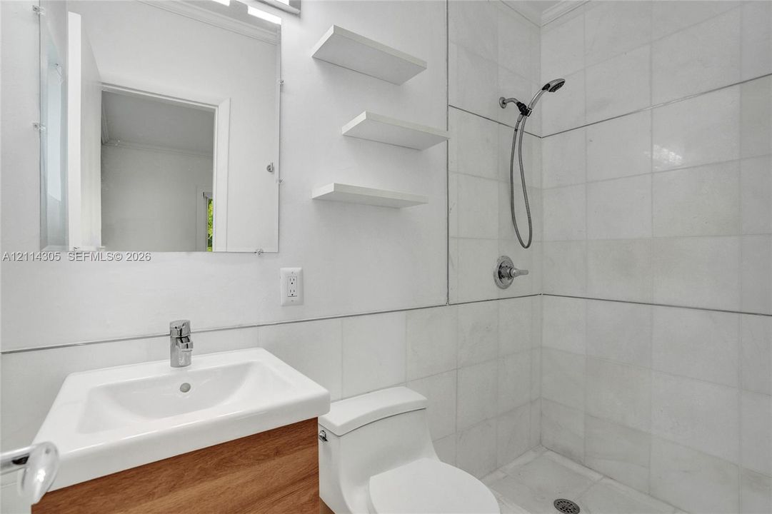 Bathroom featuring a wood-finish vanity with an integrated sink, wall-mounted shelving, a walk-in shower with tiled surround, and a handheld shower fixture