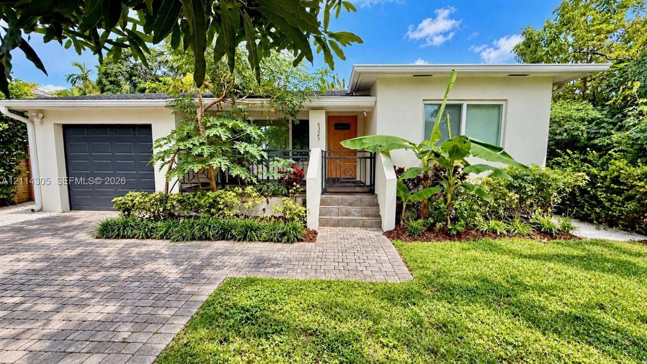 Mid-century modern exterior featuring a smooth stucco facade, a natural wood entry door, a paver stone driveway, a covered front porch, and an attached single-car garage
