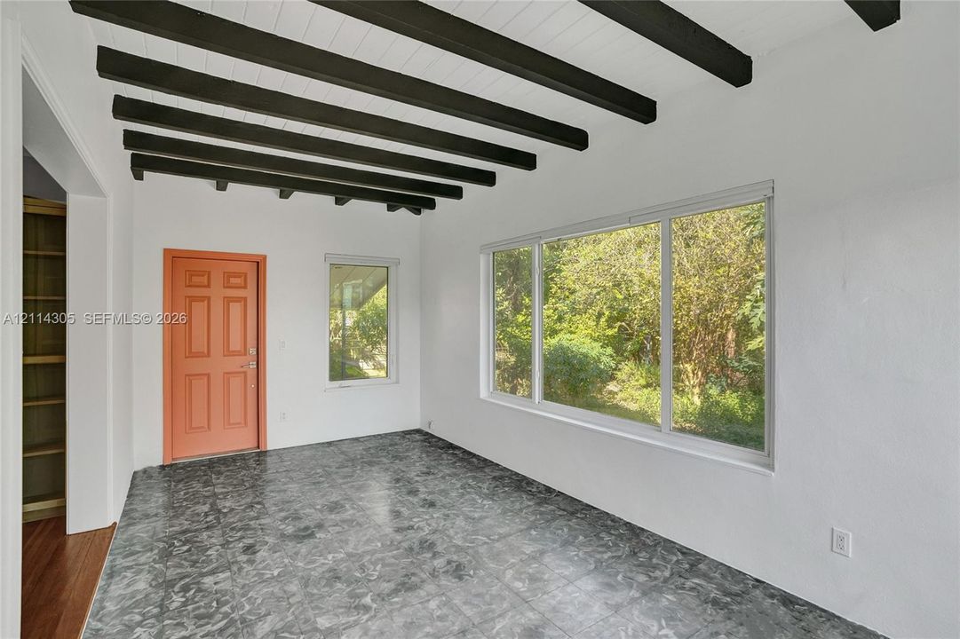 Exposed dark-finish ceiling beams, large picture window, stone-patterned tile flooring, integrated wood shelving, and exterior-facing door