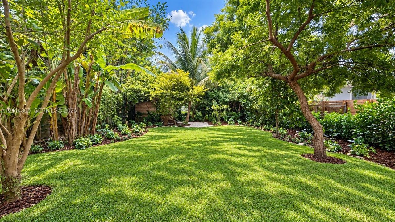 Expansive backyard featuring a manicured lawn, mature trees, dense tropical landscaping, and organic mulch garden beds