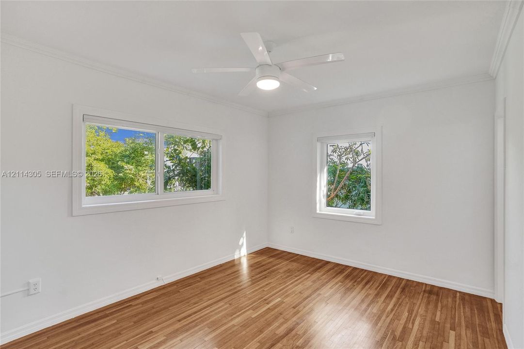 Room featuring wood-finish flooring, white walls, two windows with exterior greenery, and a white ceiling fan with integrated lighting