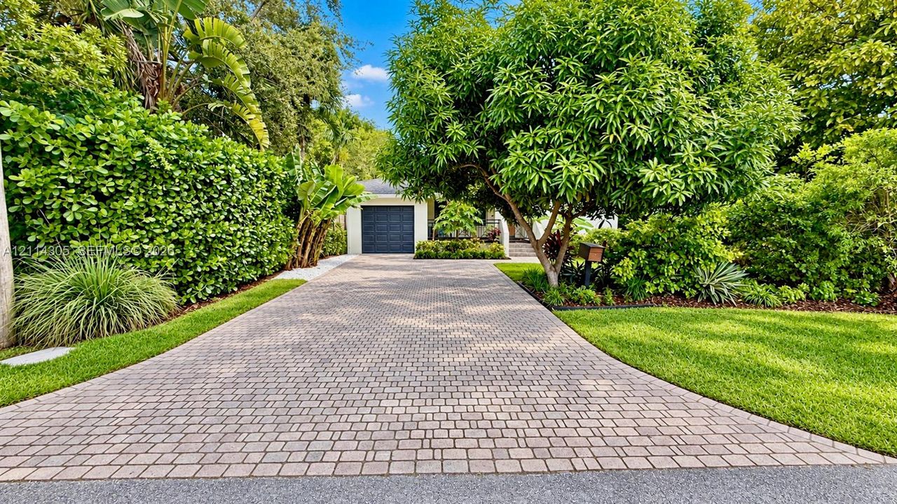 Paver driveway leading to a single-car garage, framed by mature trees, privacy hedges, and maintained lawn areas