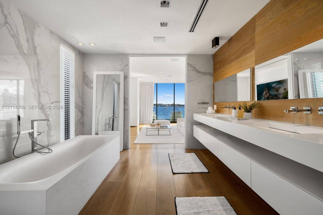 Soaking tub with handheld shower fixture, stone-surround wall detailing, floating dual vanity with integrated sinks, wood-finish wall paneling, and wood-finish flooring
