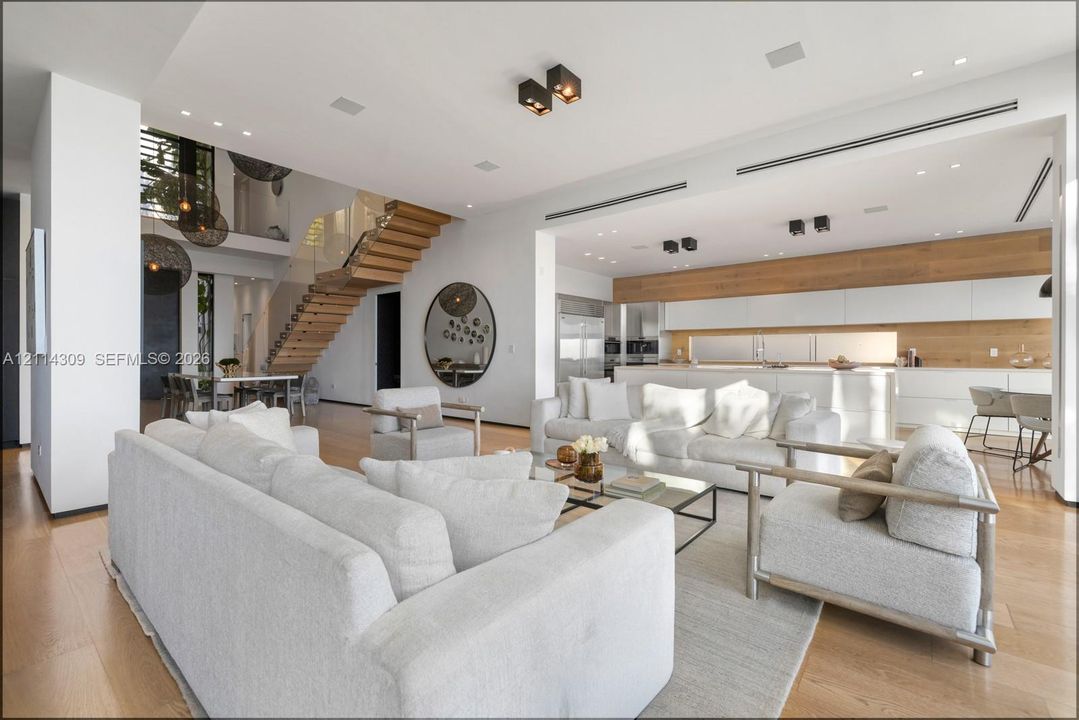 Open-concept interior featuring wood-finish flooring, a floating wood staircase with a glass railing, minimalist white cabinetry, integrated recessed lighting, and expansive ceiling heights