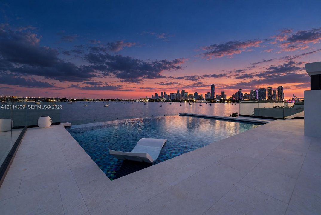 Infinity-edge swimming pool featuring a submerged lounging ledge, surrounded by expansive light-toned stone decking and transparent glass railings