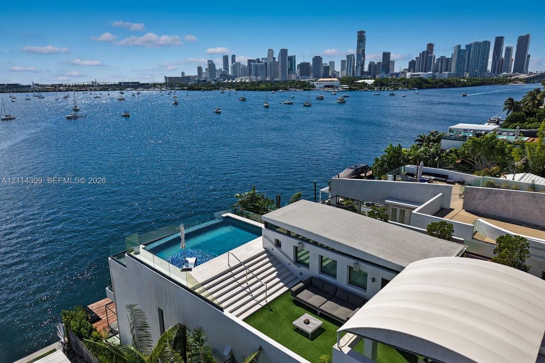 Waterfront terrace featuring an infinity-edge pool, glass railing system, integrated staircase, synthetic lawn surface, and retractable shade structure