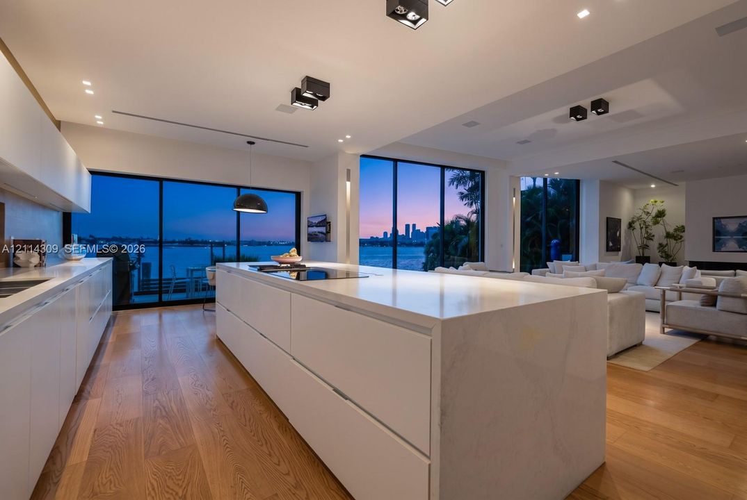 Contemporary kitchen featuring a large waterfall-edge stone island, flat-panel cabinetry, wood-finish flooring, floor-to-ceiling glass sliding doors, and recessed black architectural lighting