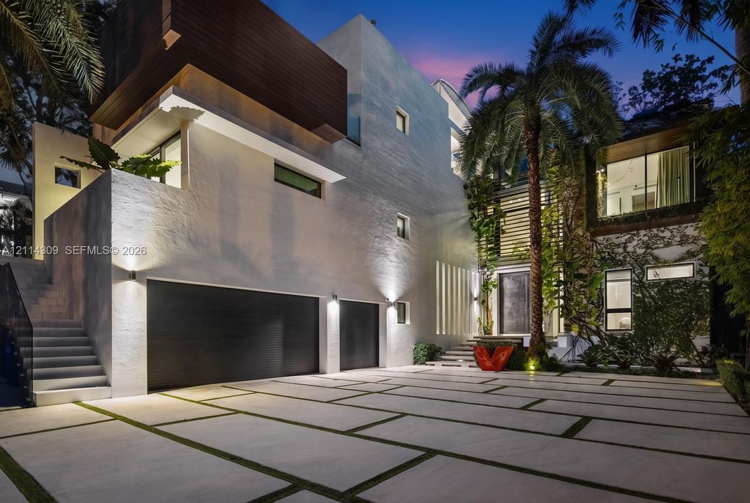 Modern architectural exterior featuring a textured white facade with wood-finished accents, multi-car garage, concrete paver driveway with integrated greenery, and custom vertical slat detailing