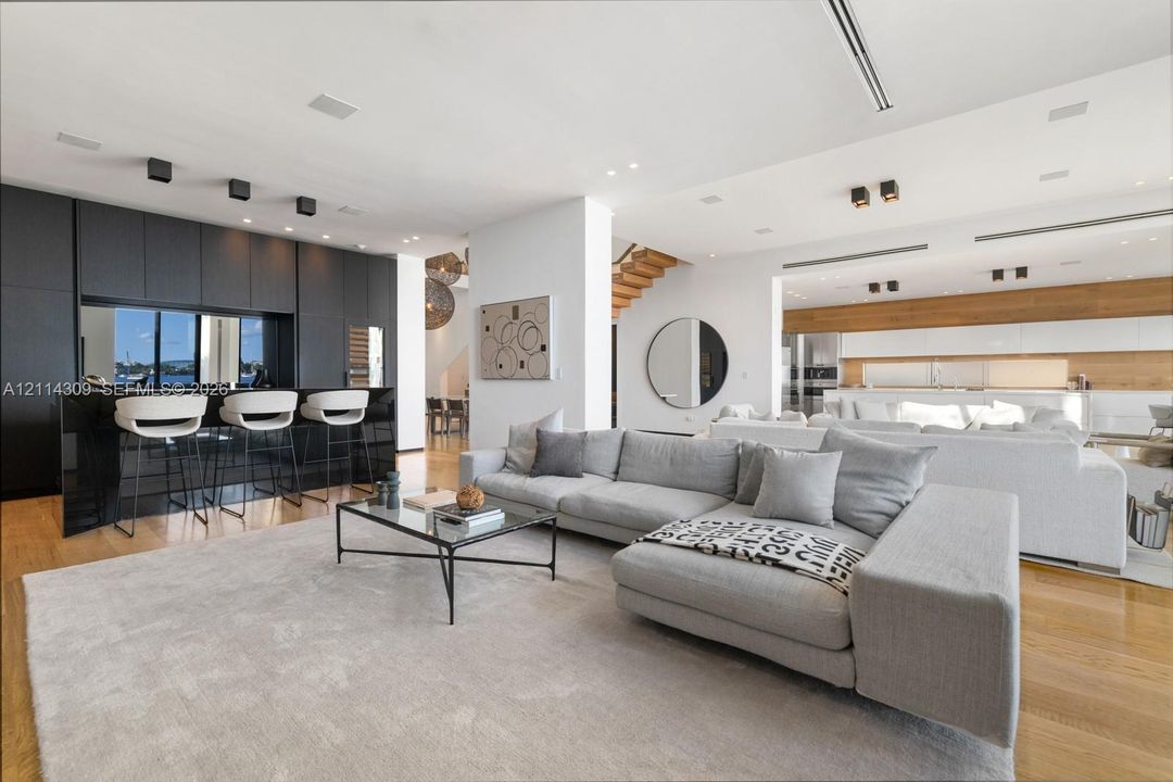 Open-concept living space featuring wood-finish flooring, custom matte black cabinetry, integrated bar seating, recessed ceiling lighting, and wooden stair detailing