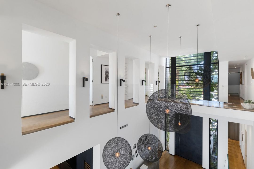 Double-height interior featuring wide-plank wood-finish flooring, custom spherical wire pendant lighting, black wall-mounted fixtures, and floor-to-ceiling multi-paned windows