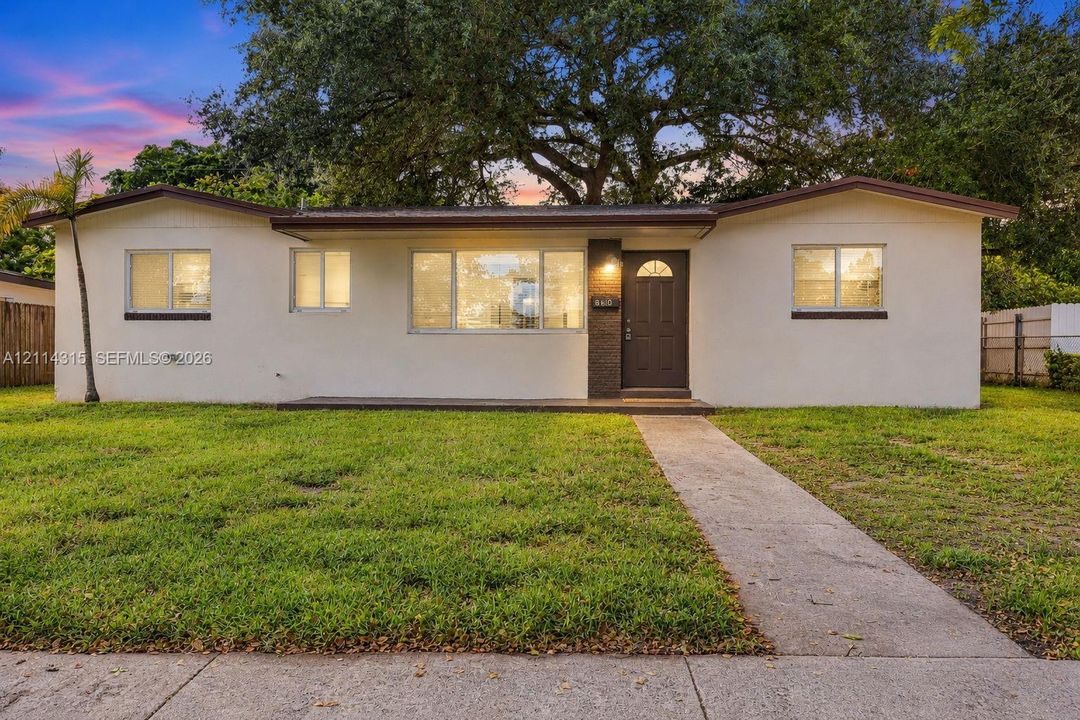 Single story exterior featuring a light-toned stucco facade, dark-finish entry door, low-pitch roofline, expansive front lawn, and mature canopy trees