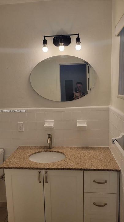 Vanity featuring a stone-composite countertop, undermount sink, white cabinetry with metallic hardware, oval mirror, and a three-light fixture