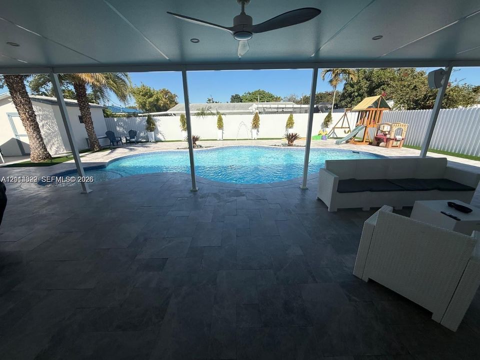 Covered patio with stone-finish tile flooring and ceiling fan, featuring a pool and a privacy wall