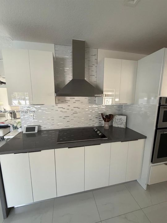 Modern kitchen featuring white flat-panel cabinetry, dark solid-surface countertops, a stainless steel chimney-style range hood, a glass-tile backsplash, and built-in double wall ovens