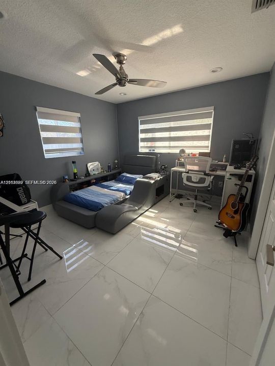 Room featuring large-format white tile flooring, neutral grey walls, dual-layered roller window shades, and a ceiling fan with integrated lighting