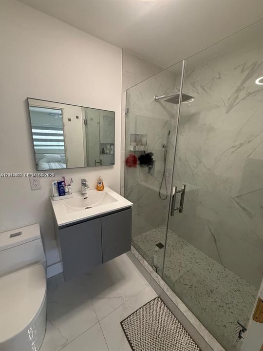 Floating vanity with a rectangular basin, frameless glass-enclosed shower, wall-mounted rain shower head, large-format tile flooring, and neutral-tone wall surfacing