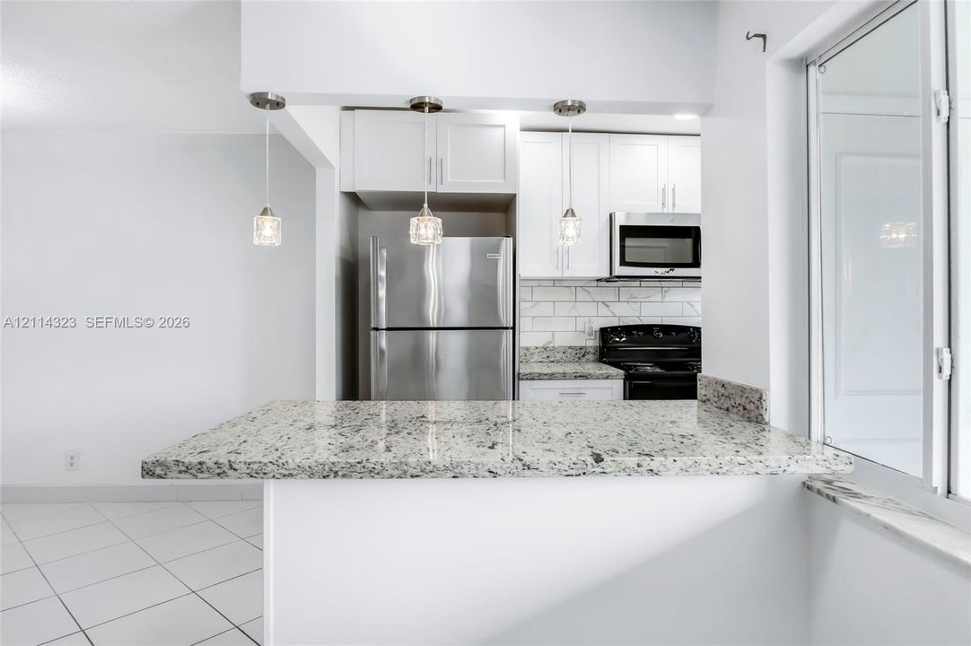 Kitchen featuring a peninsula with stone-finish countertop, three glass pendant lights, white shaker-style cabinetry, stainless steel appliances, and subway tile backsplash