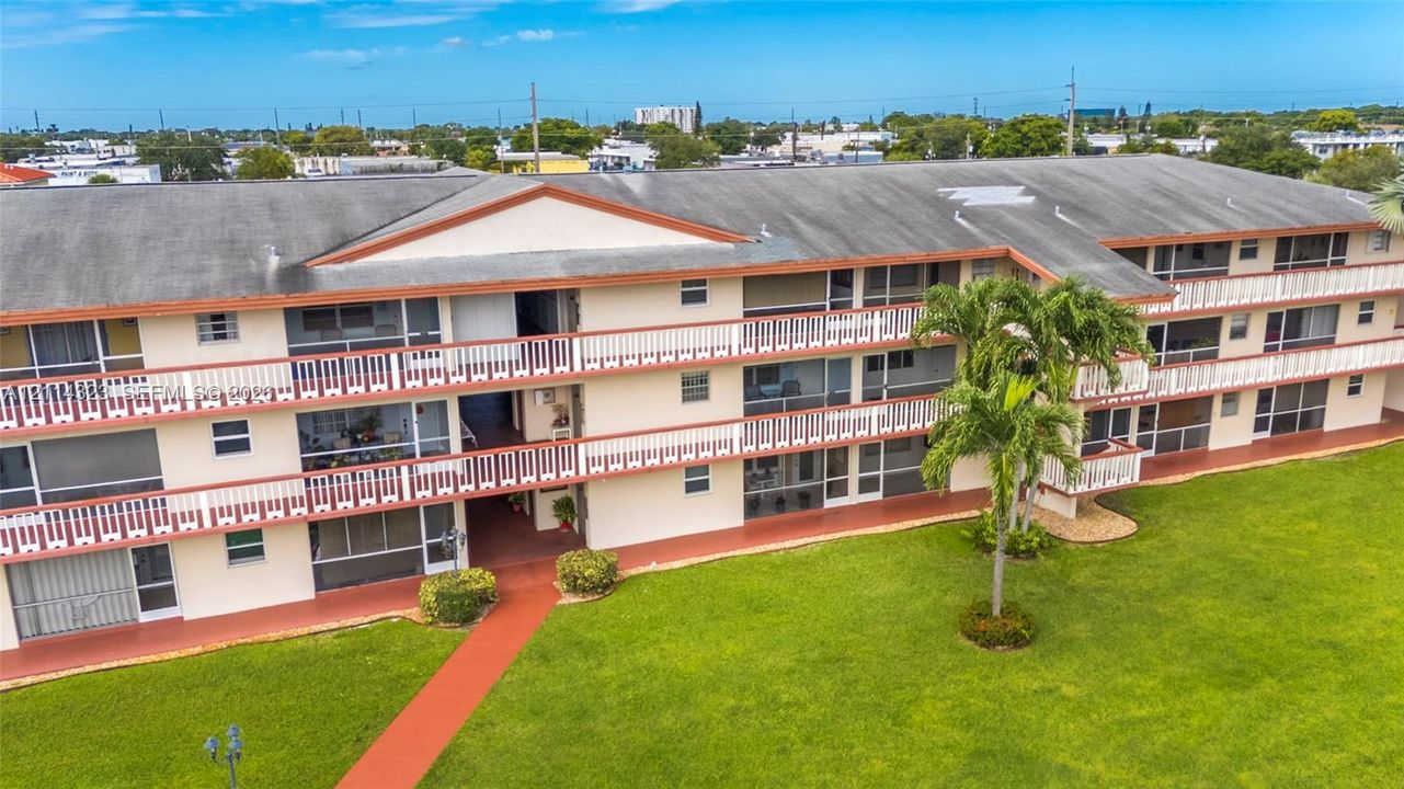 Multi-story residential building featuring covered private balconies with railings, exterior walkways, a gabled roofline, and expansive surrounding lawn areas with mature palms
