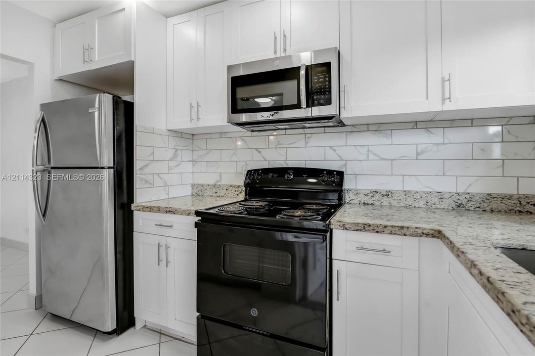 Kitchen featuring white shaker-style cabinetry, stone-finish countertops, subway tile backsplash, stainless steel microwave, and stone-tile flooring