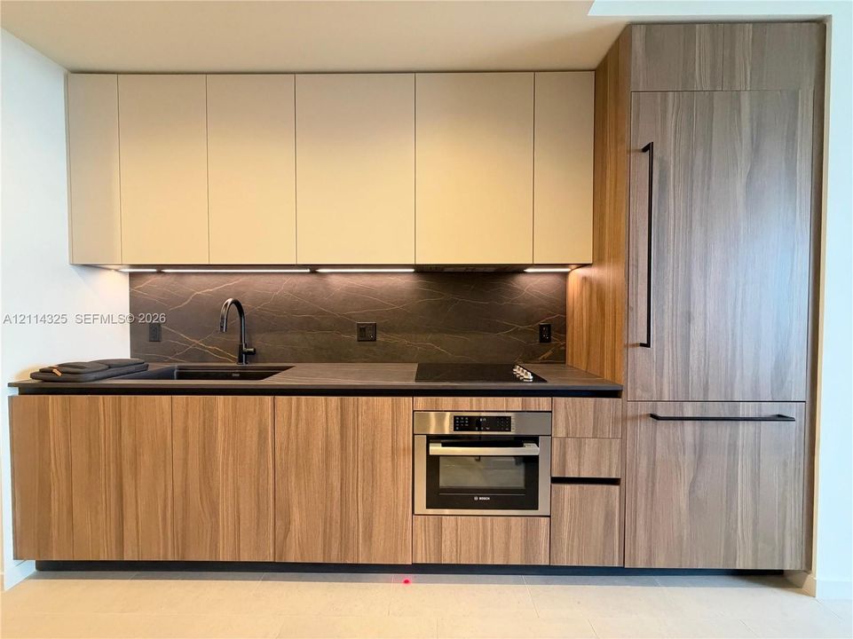 Kitchen featuring wood-finish cabinetry, integrated panel-ready refrigerator, matte black faucet, dark stone-finish backsplash, and built-in electric cooktop with stainless steel oven
