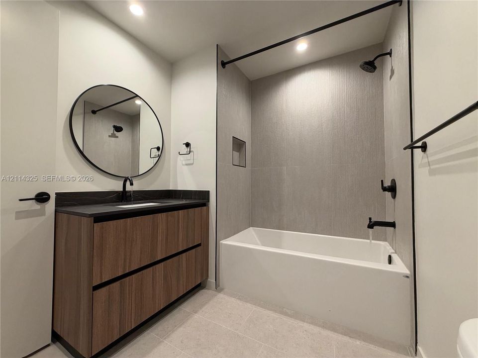 Bathroom featuring a wood-finish floating vanity, dark countertop, round mirror, and a tub-shower combination with matte black fixtures