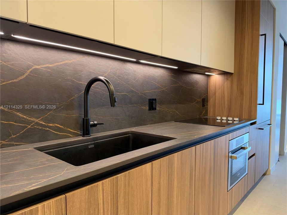 Sleek kitchen featuring flat-panel wood-finish cabinetry, dark stone-finish countertops and backsplash, integrated under-cabinet lighting, a matte black high-arc faucet with an undermount sink, and a built-in wall oven and cooktop