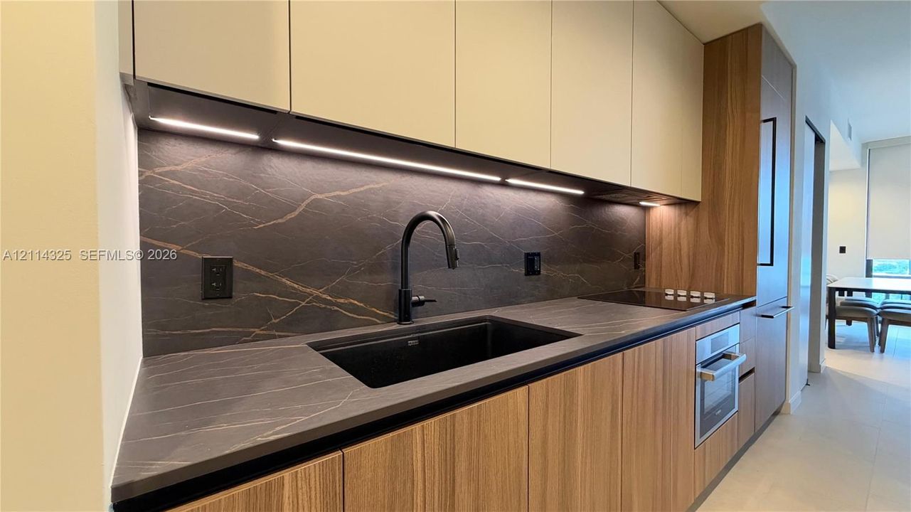 Modern kitchen featuring wood-finish lower cabinetry, matte white upper cabinets, dark stone-finish countertops and backsplash, integrated under-cabinet lighting, and a black gooseneck faucet