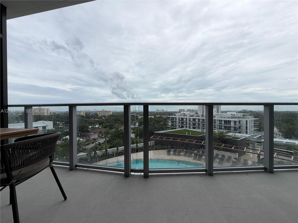 Covered balcony with a glass railing, concrete flooring, and surrounding area scenery including a swimming pool