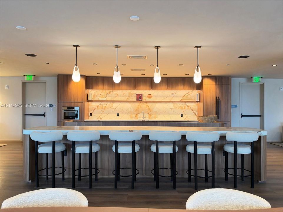 Custom kitchen featuring wood-finish cabinetry, a full-height stone backsplash, an expansive island with a waterfall countertop, integrated stainless steel appliances, and four pendant light fixtures