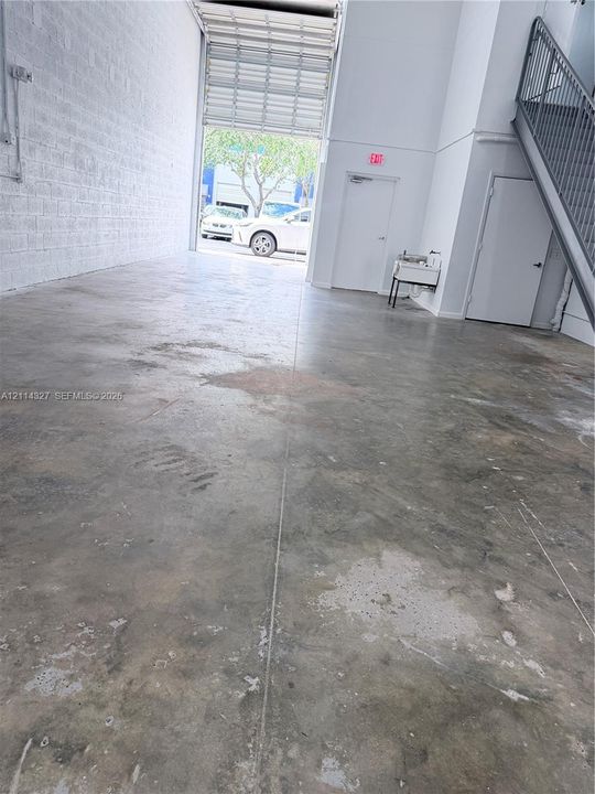 High-ceiling industrial interior featuring a large roll-up bay door, concrete flooring, exposed masonry wall, and a steel staircase with railing