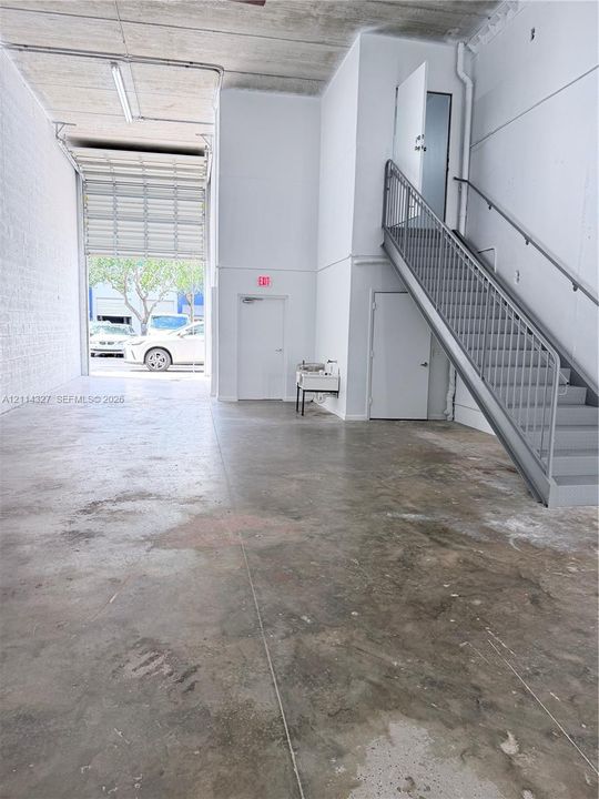 Industrial space featuring high ceilings, concrete flooring, a roll-up bay door, a metal staircase with handrails, and a wall-mounted utility sink