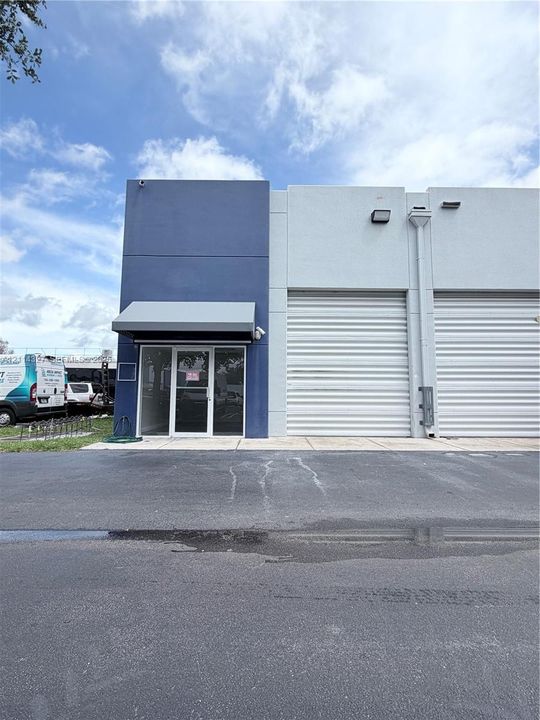 Industrial exterior featuring dual roll-up loading doors, a glass-paneled pedestrian entrance, metal awning, and exterior lighting fixtures