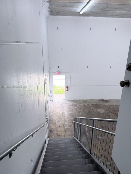 Industrial staircase featuring metal treads, a tubular steel handrail, and a vertical metal balustrade, opening to a concrete floor area with high-clearance walls and an exit door