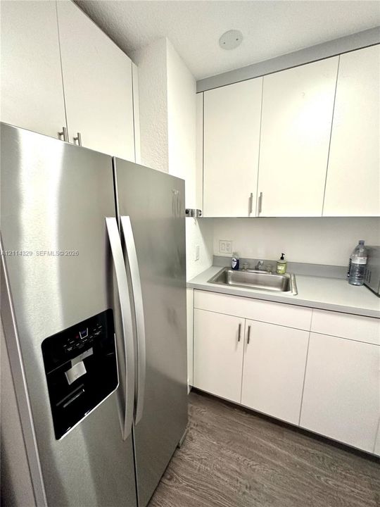 Stainless steel side-by-side refrigerator, white cabinetry with brushed metal hardware, solid-surface countertop, and integrated sink with faucet fixture, complemented by wood-finish flooring